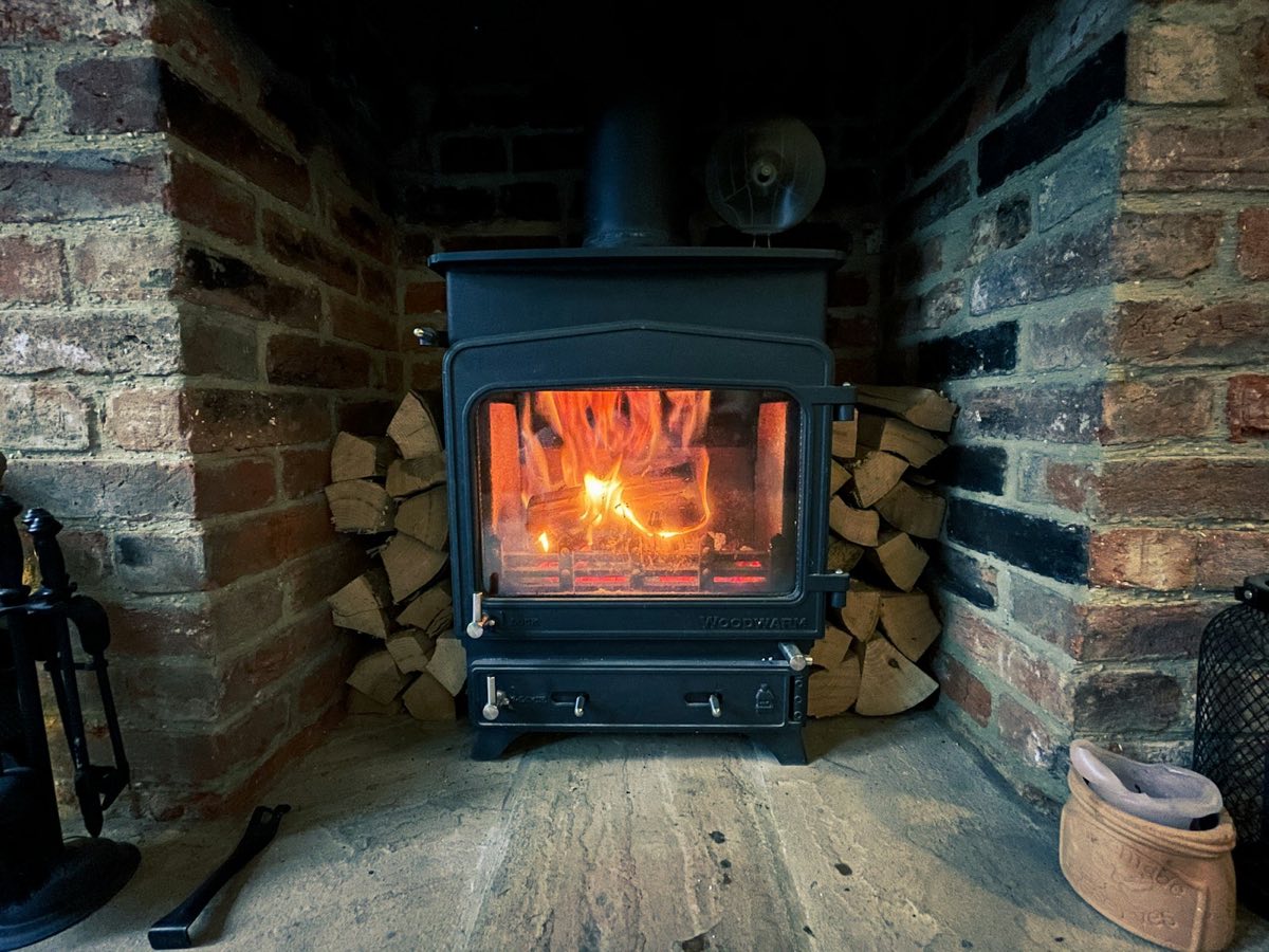 A wood-burning stove alight in a brick fireplace, with split logs stacked either side